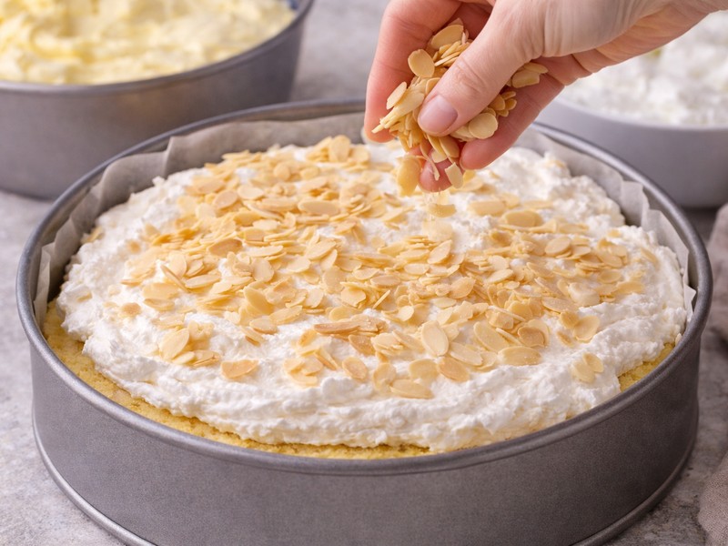 Eine Hand streut Mandelblättchen über eine Schicht weißer Creme auf einem Biskuitboden in einer Backform.